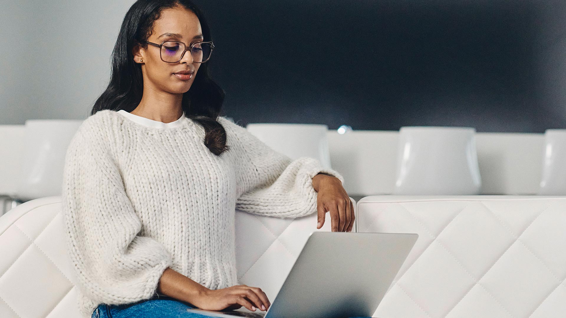 Dark-haired woman with dark glasses and a white sweater looks into a laptop.