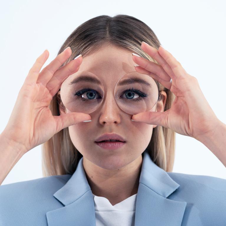 A young blonde woman holds lenses in front of her eyes to show the fisheye effect caused by thick glasses.