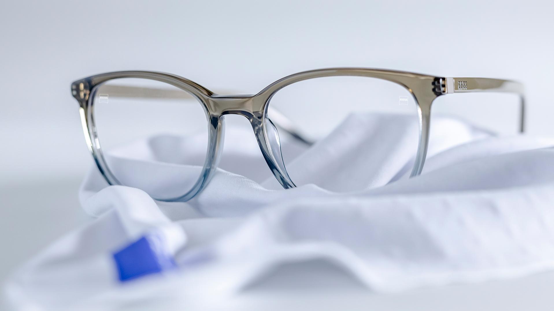 A pair of glasses with grey-blue frames and ZEISS lenses with DuraVision® coating lies on a white microfibre cloth.