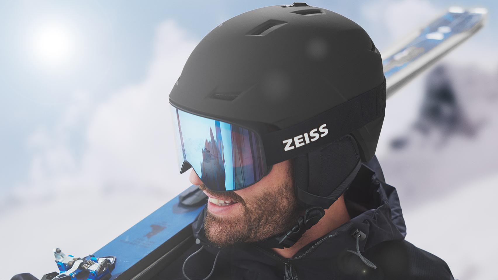 A man wears a black helmet with blue mirrored ZEISS ski goggles, has skis over the right shoulder.