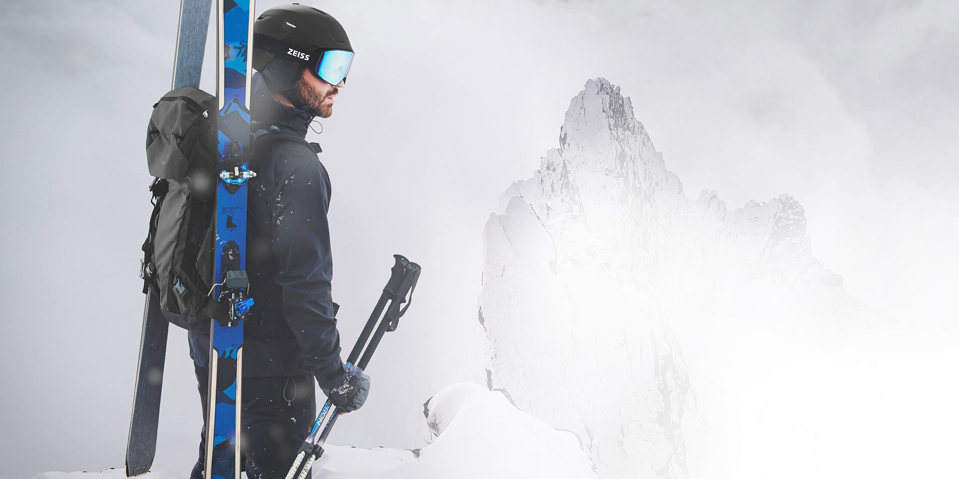 A man wears a black helmet with blue mirrored ZEISS ski goggles, has skis and a backpack on his back and holds the ski poles in his right hand. He is standing on a mountain top.