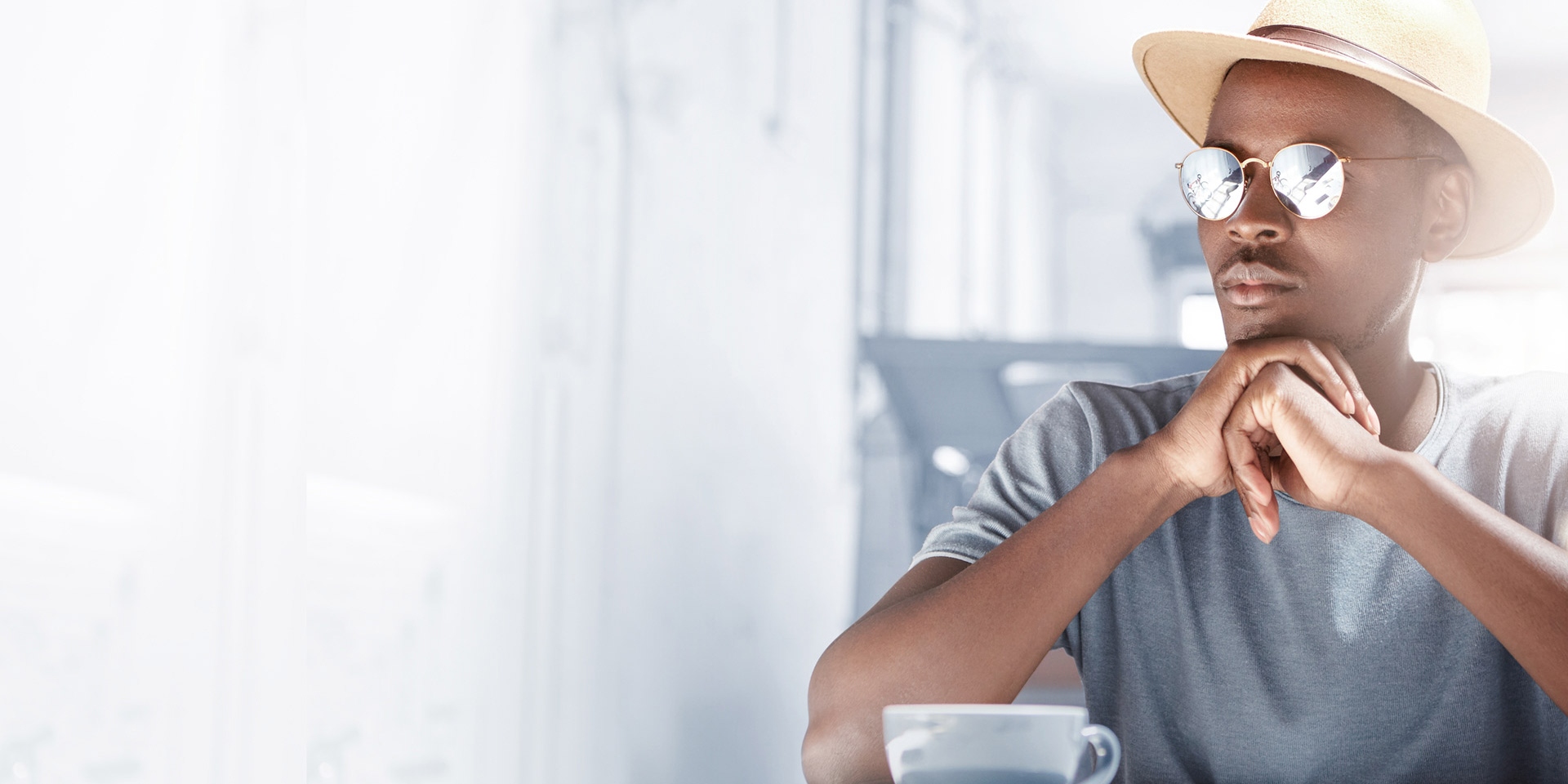 Indoor shot of attractive young Afro American man dressed in fashionable clothing and eyewear waiting for his girlfriend for lunch, sitting at cafe table with mug and cell phone, having pensive look